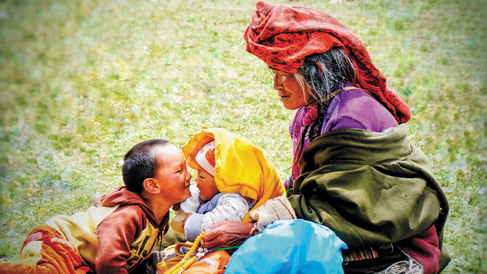 An elderly Mongolian woman holding a young baby face to face with a young Mongolian boy on the grass in vivid clothing