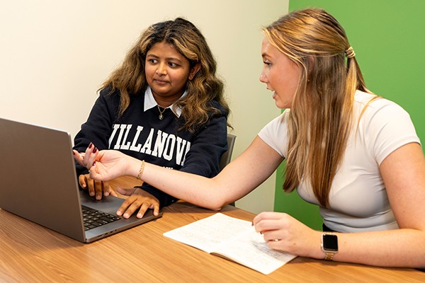 students sitting together at a table, one has a laptop, the other is writing with a pen and notepad