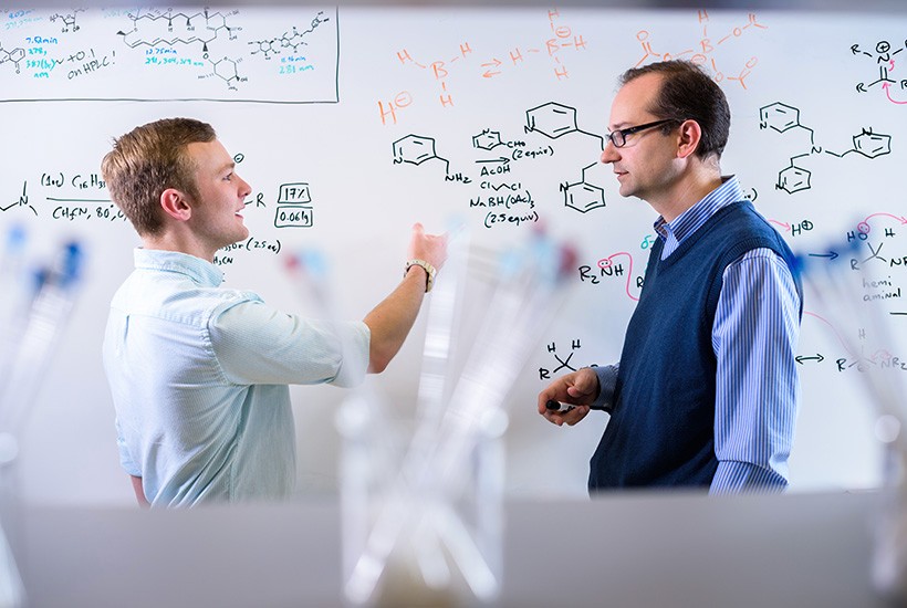 image of student and professor discussing chemical equations at a whiteboard.