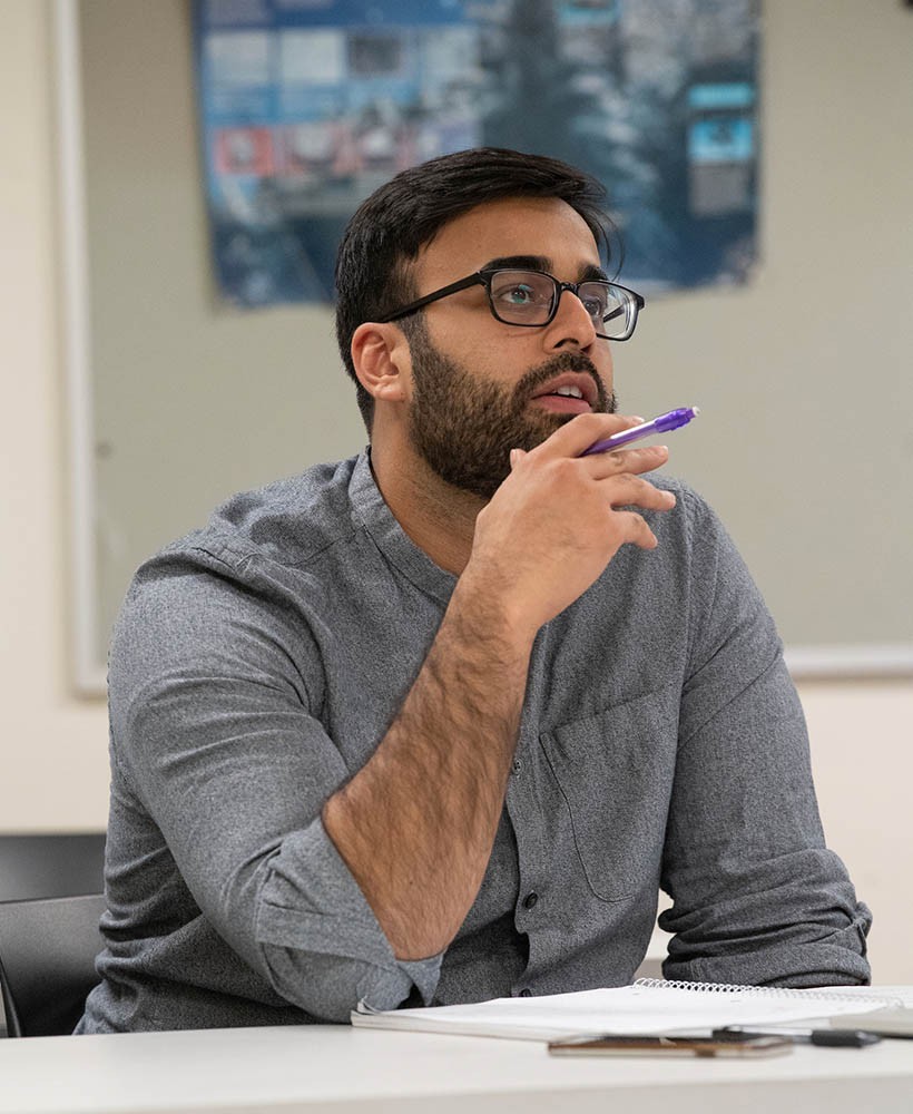 A student sits at a desk looking thoughtfully toward the front of the classroom.