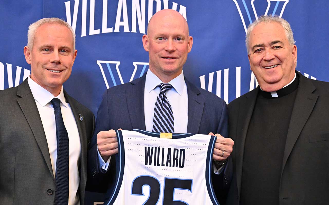 Eric Roedl, Kevin Willard, and Father Peter pose in front of a Villanova backdrop as Willard holds a Villanova jersey with his name on it.